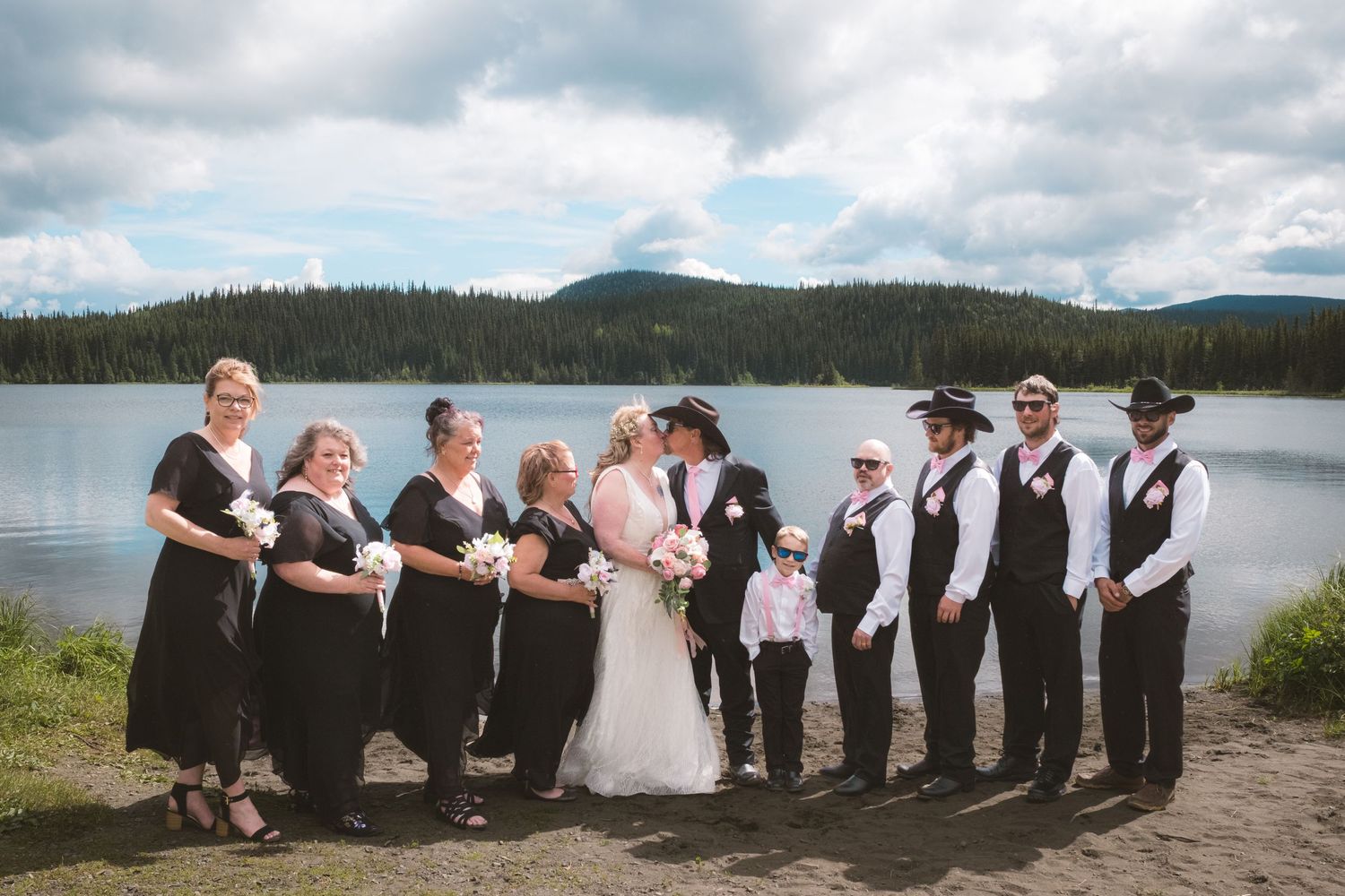 Wedding party poses by a mountain lake, bridesmaids in black dresses with groomsmen in vests and ties.