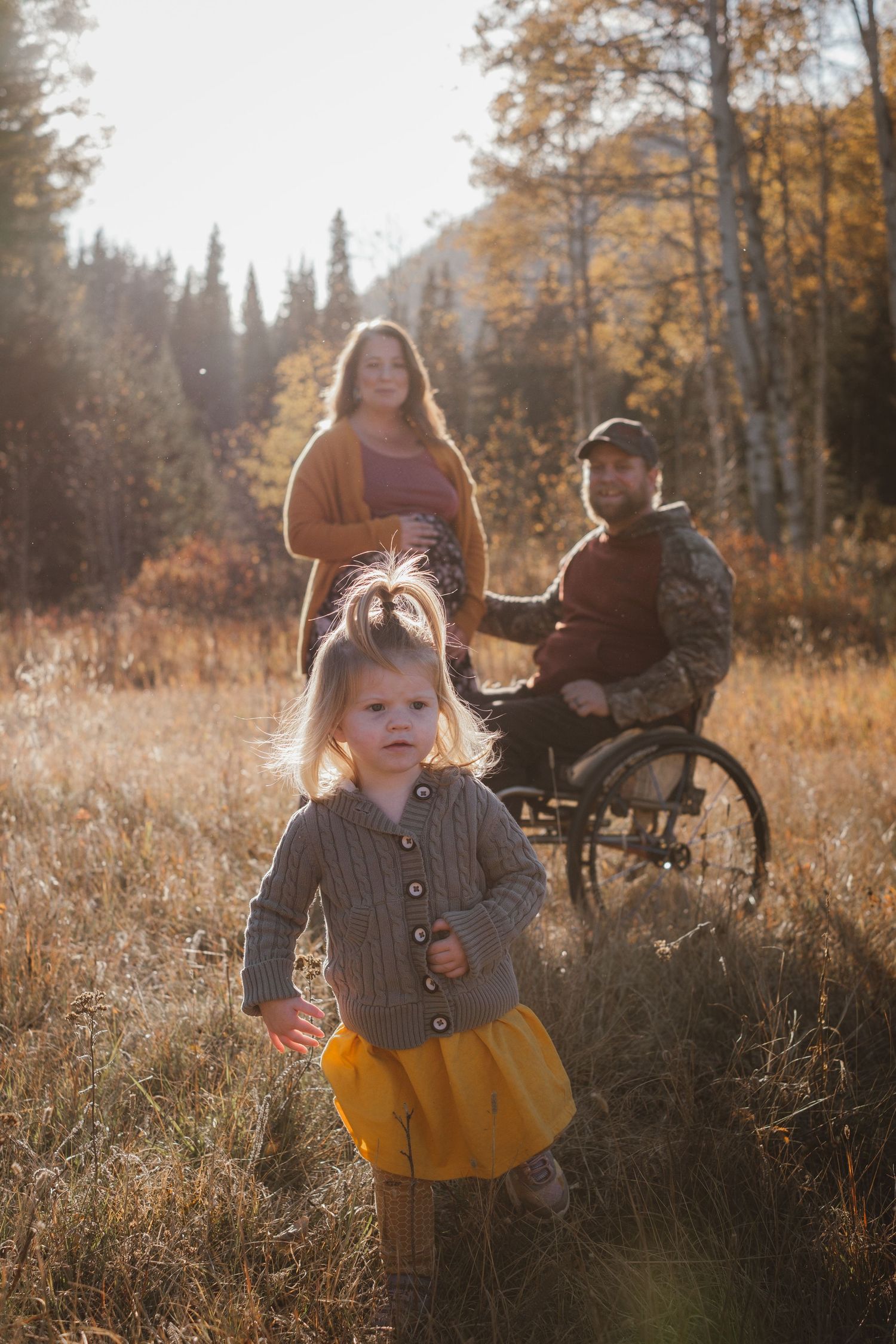 Young blonde toddler girl stands in golden autumn field with parents, father seated in wheelchair, in background.