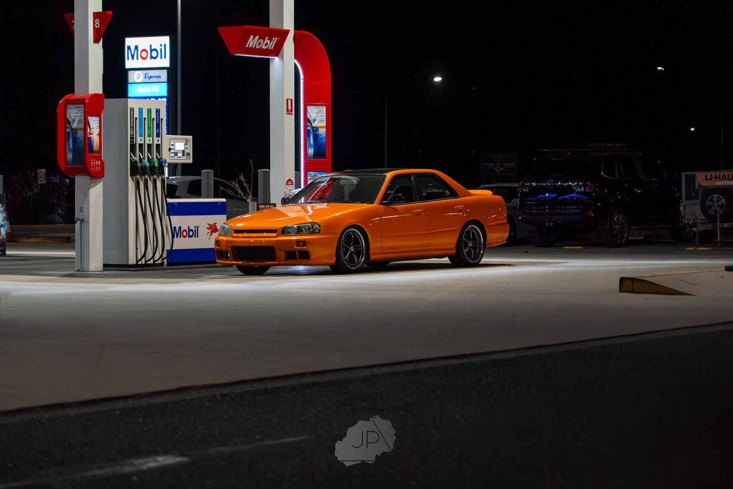 Orange sports car parked at a dimly lit gas station at night with neon lighting visible in the background.