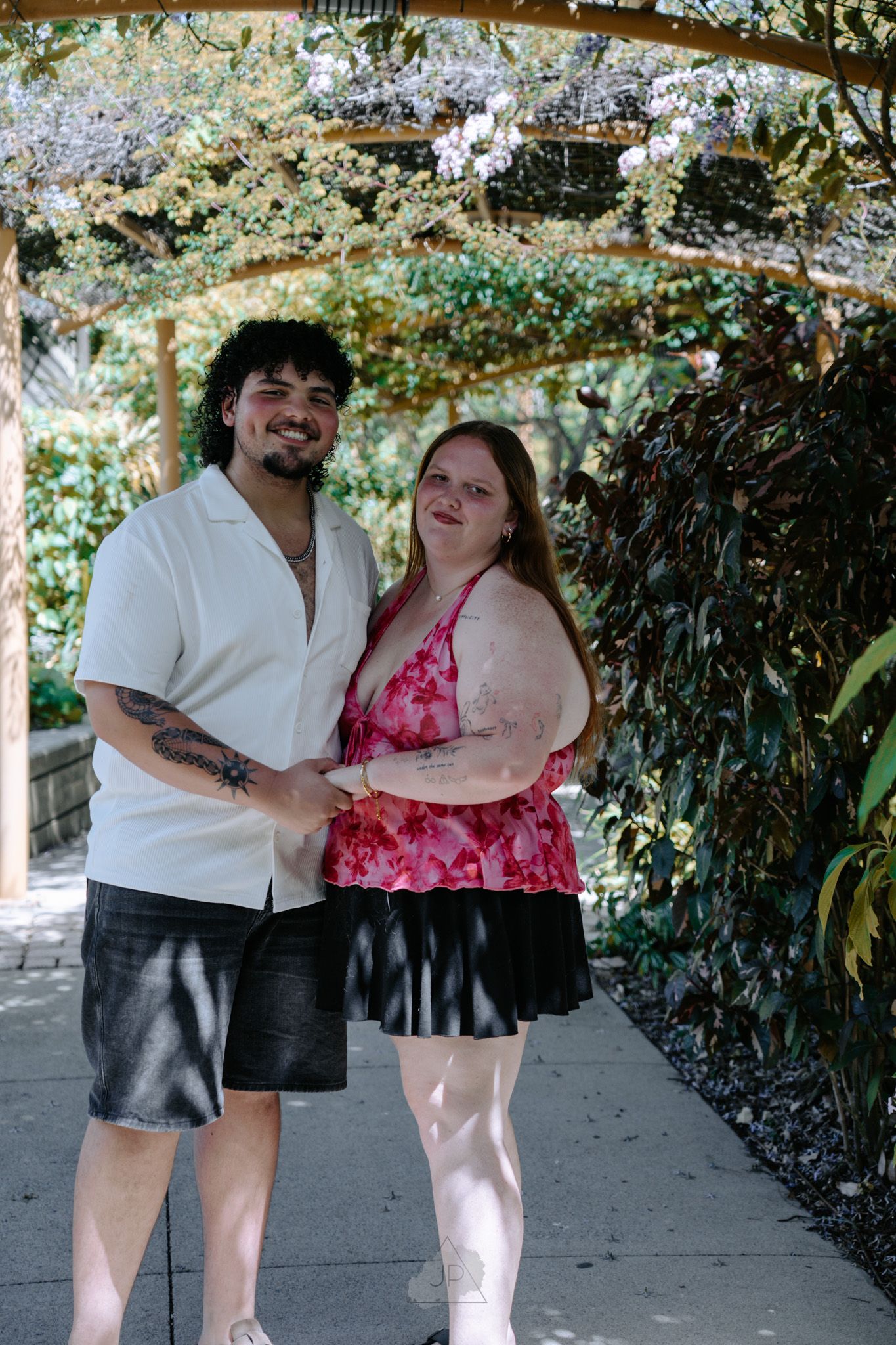 A couple poses together under blooming wisteria on a sunny garden walkway.