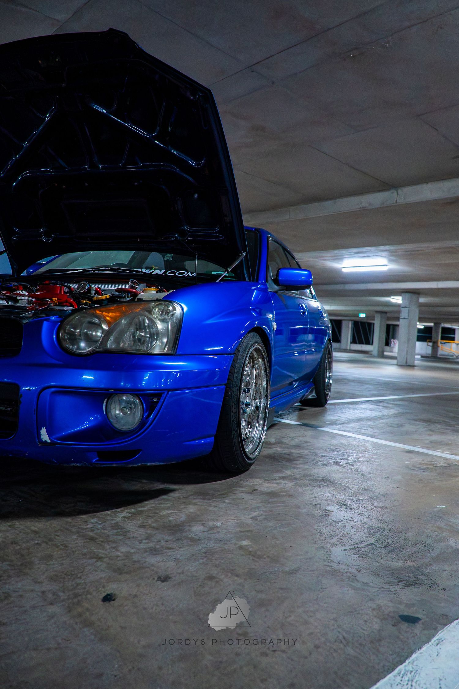 Low angle shots of modified blue Subaru WRX in parking garage showing custom wheels and front end styling elements.