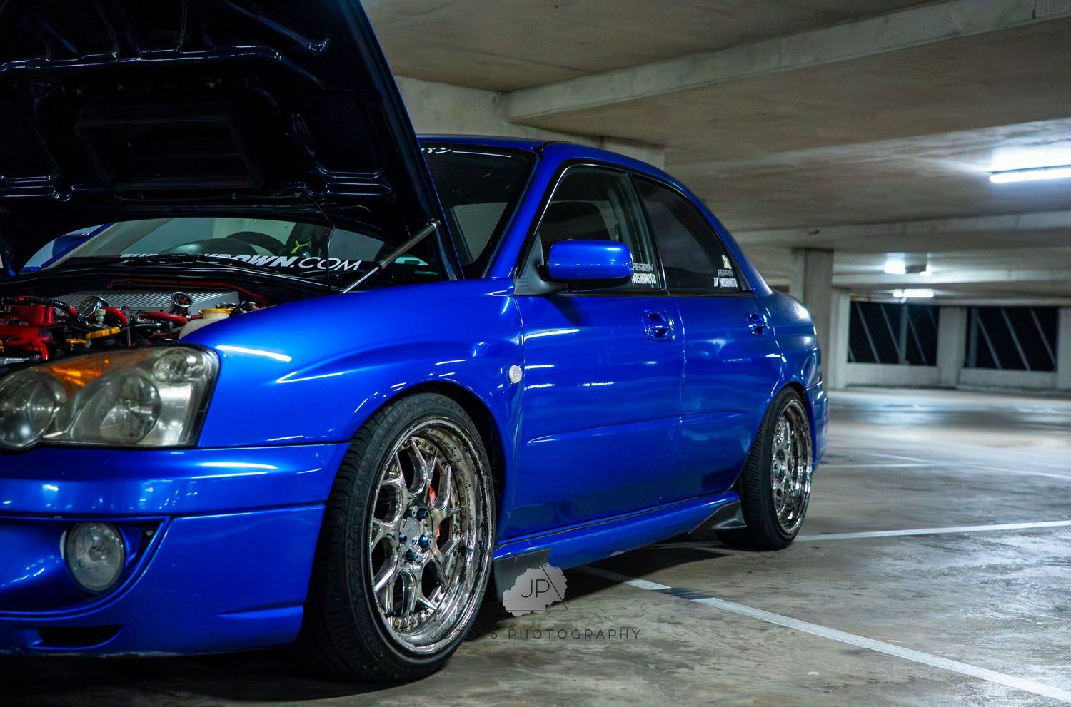 A lowered blue sports car with custom wheels sits in a parking garage with dramatic lighting.