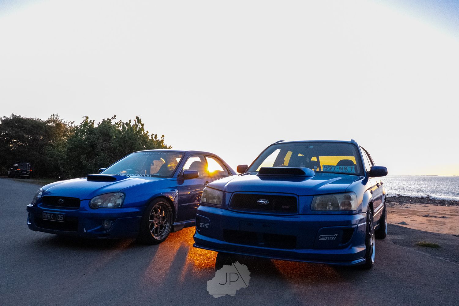 Multiple Subaru WRX STI performance cars lined up at sunset with vibrant blue paint against a scenic backdrop.