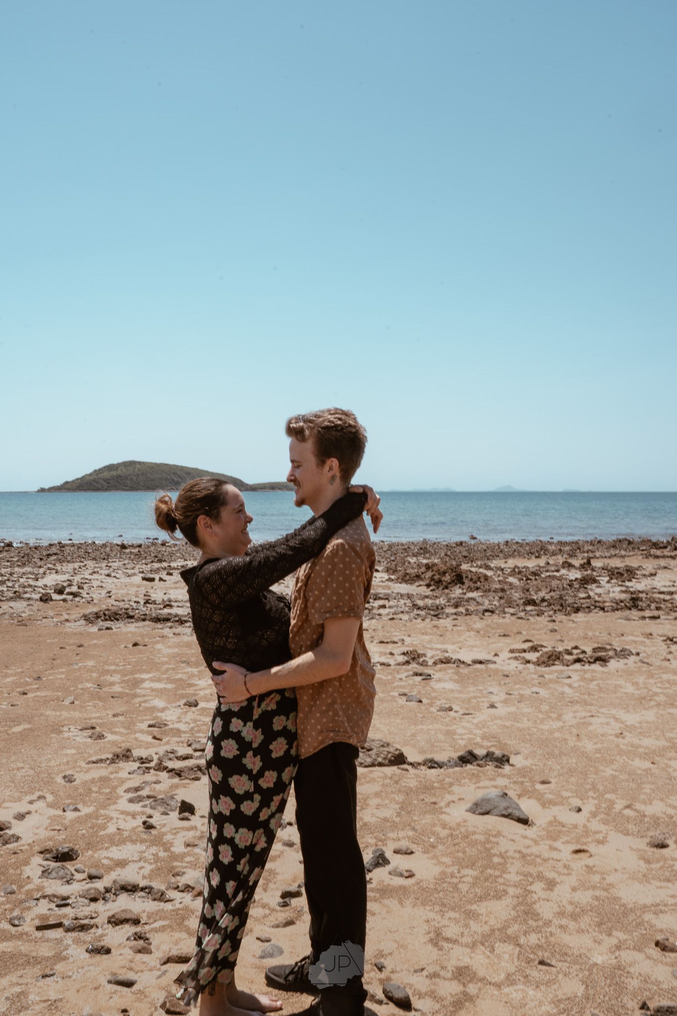 Two people embrace on a rocky beach with a distant mountain visible on the horizon.