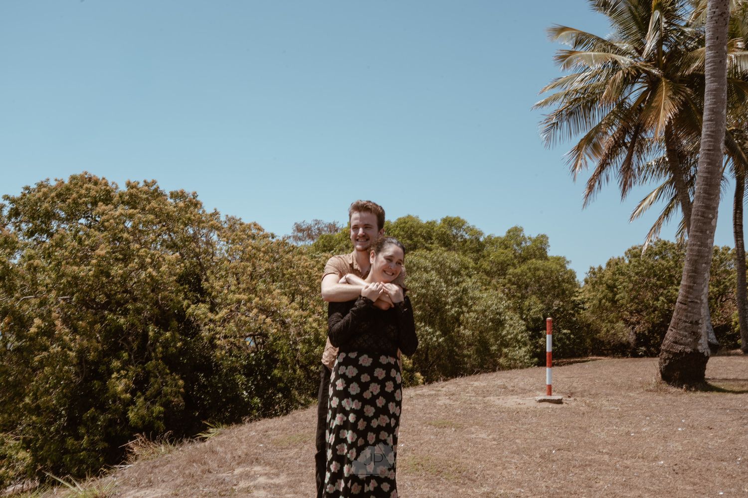 Person in floral skirt stands with crossed arms on sunny tropical path with palm trees and lush greenery.