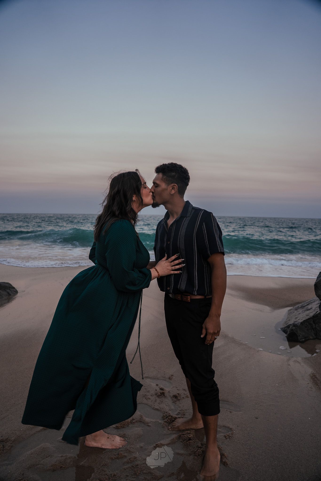 A couple shares romantic moments on a beach at sunset as waves crash against the shore.