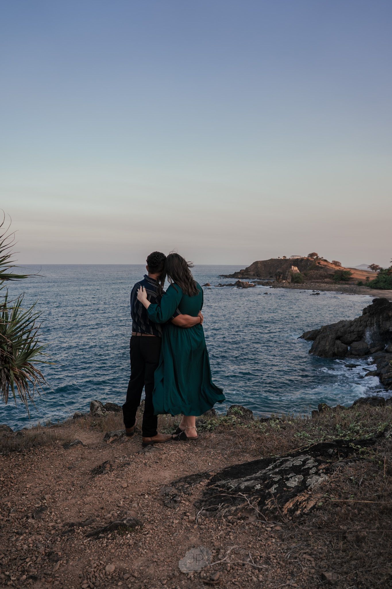 A couple shares a romantic moment on a rocky coastal cliff overlooking the ocean at sunset.