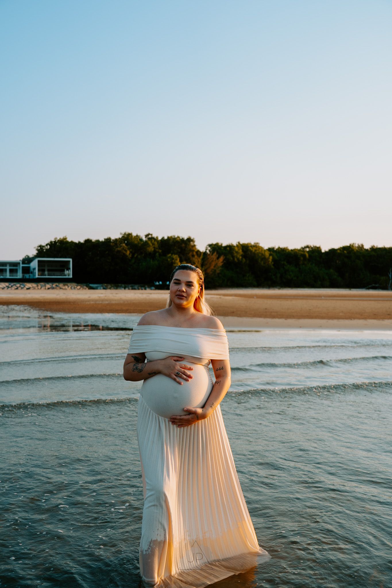 A pregnant woman in a flowing white dress stands in shallow ocean water at sunset on a peaceful beach.