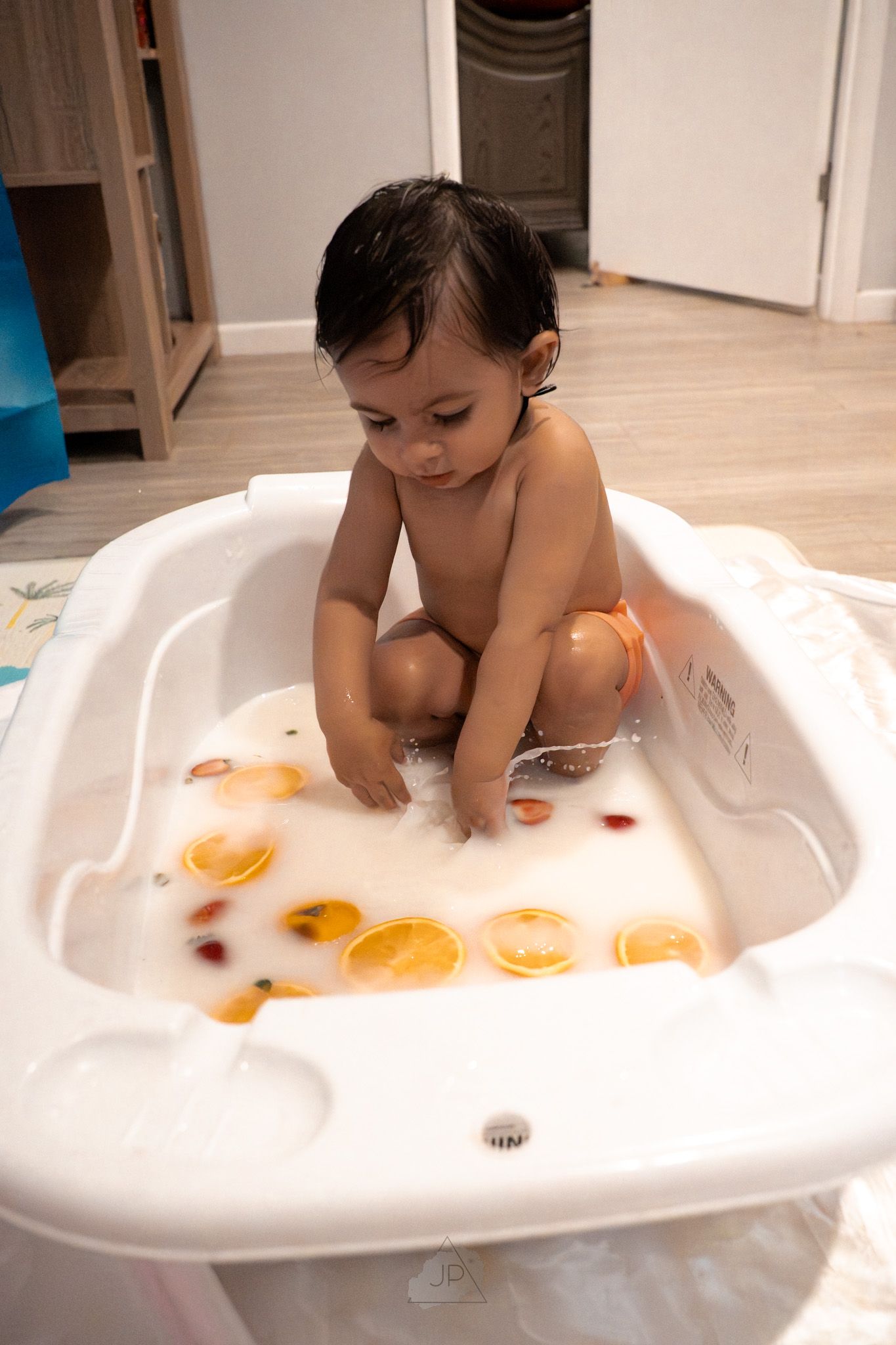 Baby playing with orange slices during bath time in white tub filled with bubbles and water.