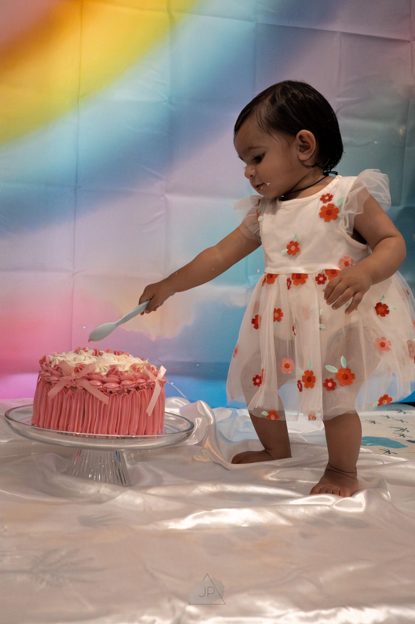 A small child in a white dress with red flowers reaches towards a pink birthday cake on a cloudy backdrop.