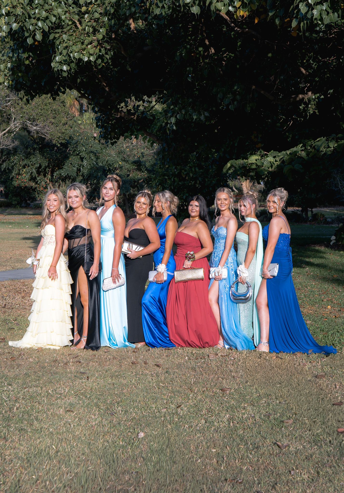 A group of women in formal dresses in shades of blue and red pose together outdoors on a grassy lawn.
