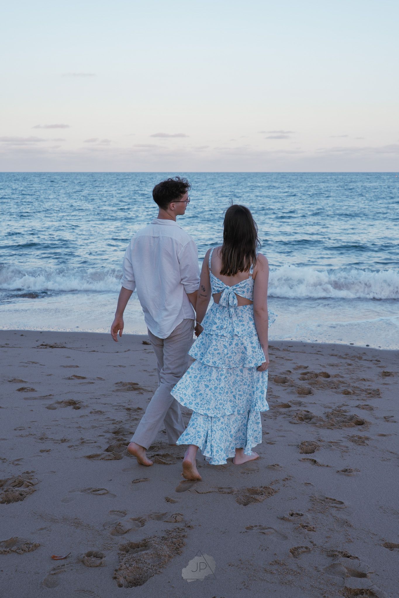 A couple in white and blue attire walks together along a sandy beach at sunset with ocean waves in the background.