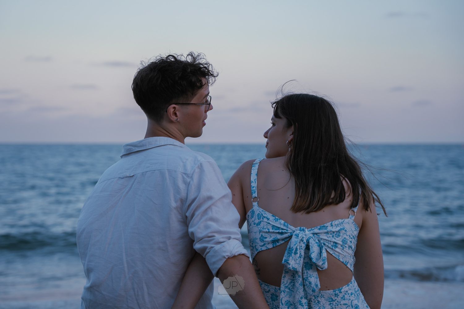 A couple in white clothing walks along a beach at sunset holding hands and embracing near the ocean.