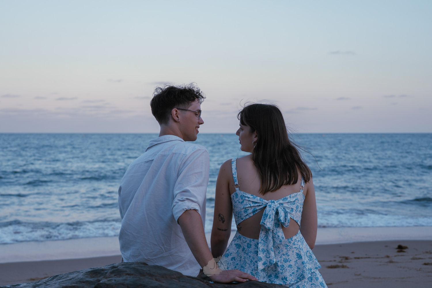 A couple sits together on a sandy beach at sunset watching the ocean waves roll in.