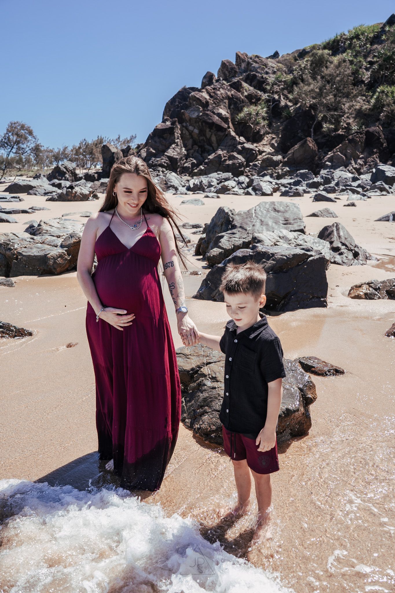 A pregnant woman in a burgundy dress and child stand on a beach with rocky cliffs and ocean waves in Hawaii.