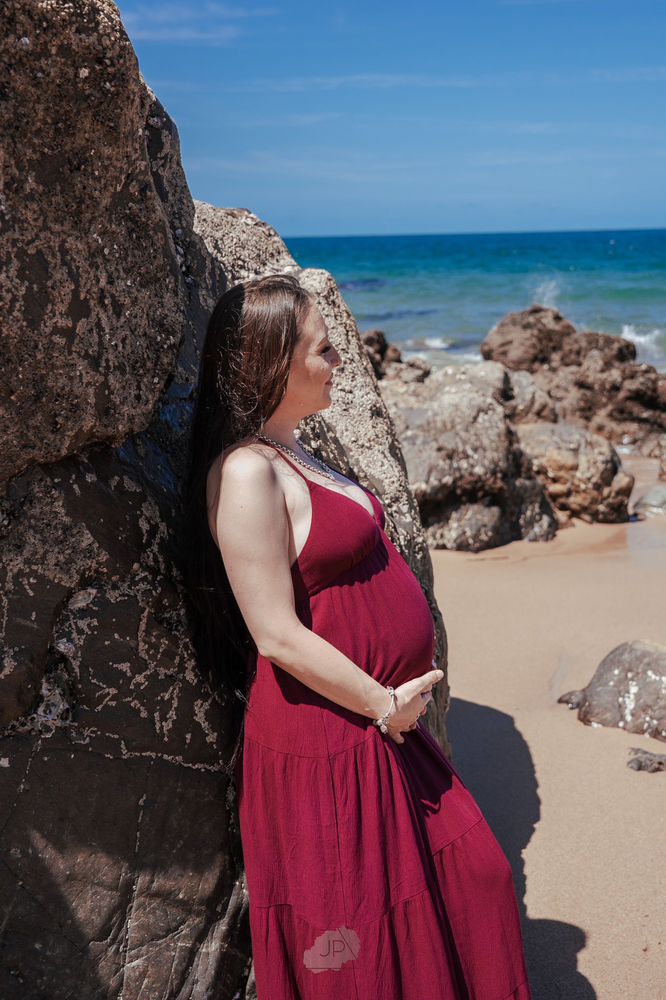 A person in a burgundy dress poses against rocky cliffs at a scenic beach with turquoise waters and blue skies.