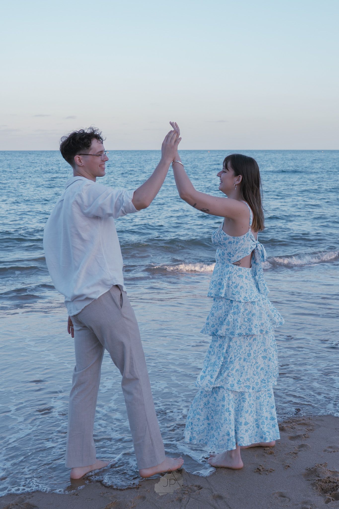 A couple in white and blue outfits playfully high-fiving on a beach at sunset with ocean waves in the background.