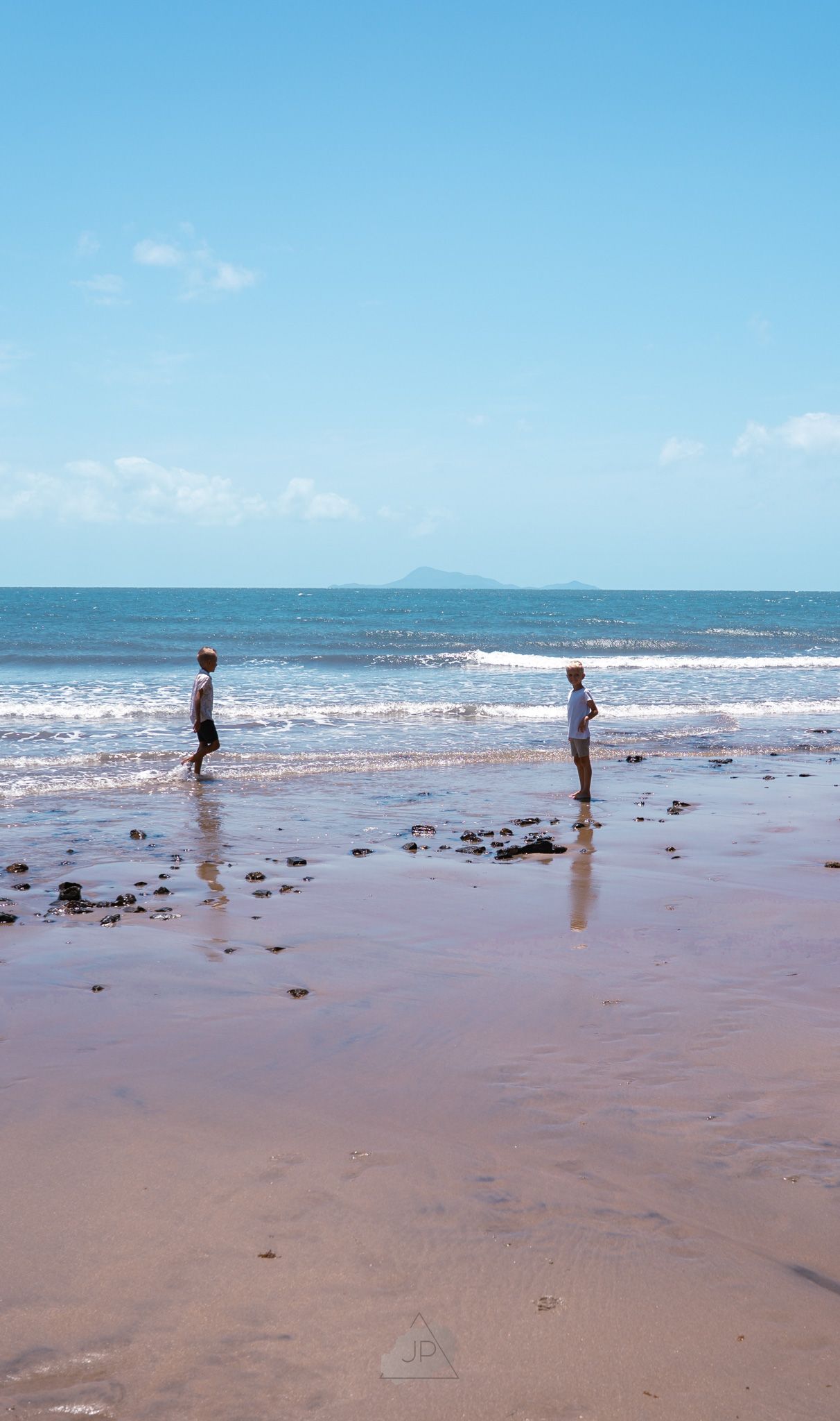 Two people stand at the edge of ocean waves on a sandy beach with mountains visible in the distance on a sunny day.