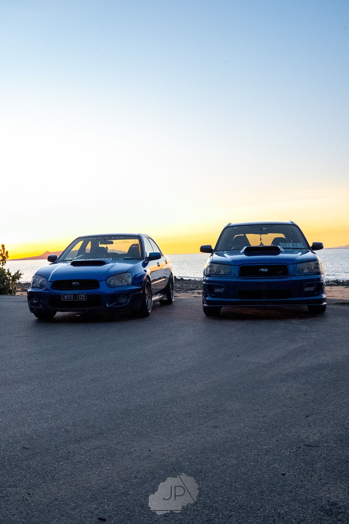 Two blue Subaru WRX cars parked at sunset against scenic coastal skyline backdrop.