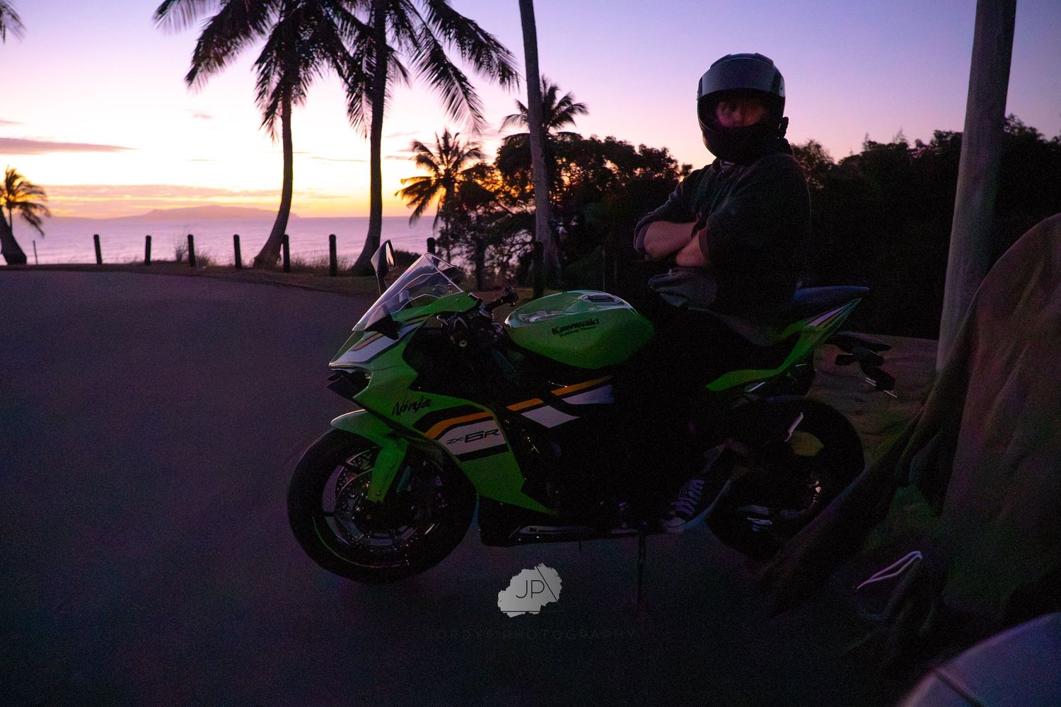A motorcyclist on a green sport bike silhouetted against palm trees and a purple sunset sky at the beach.