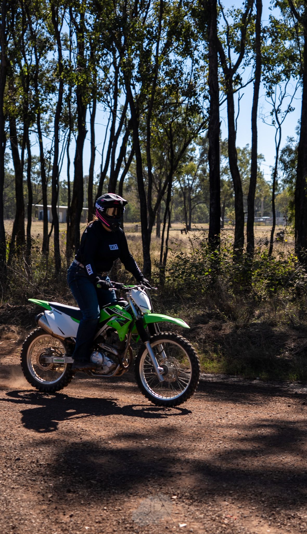 Motorcyclist riding a green dirt bike through a sunlit wooded trail on a sunny day.