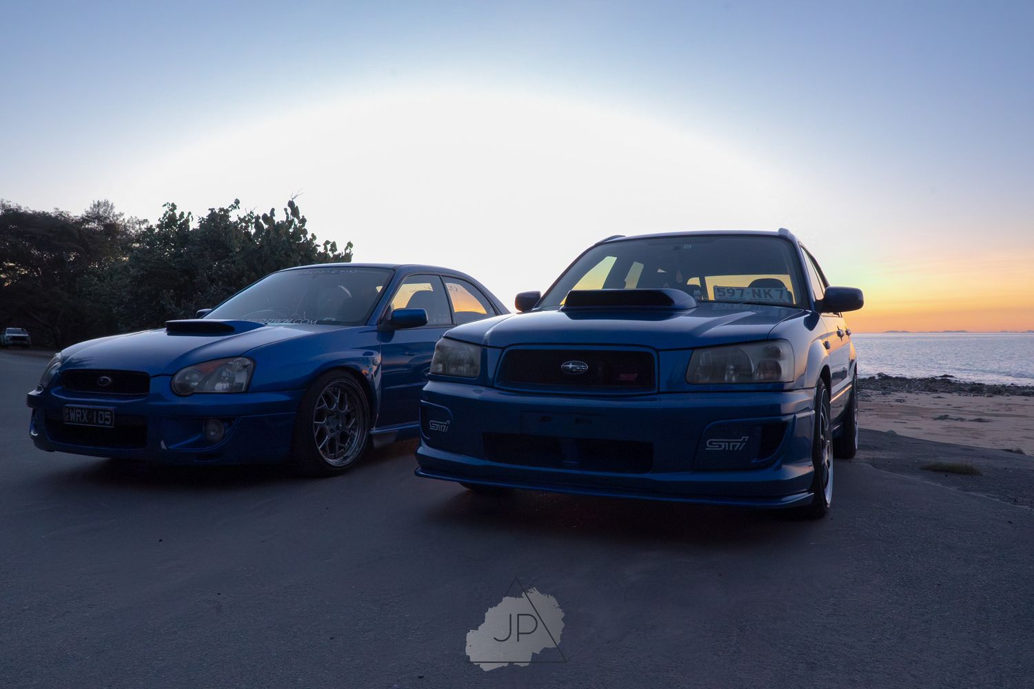 A group of blue Subaru WRX sports cars parked at sunset with scenic mountain views in the background.