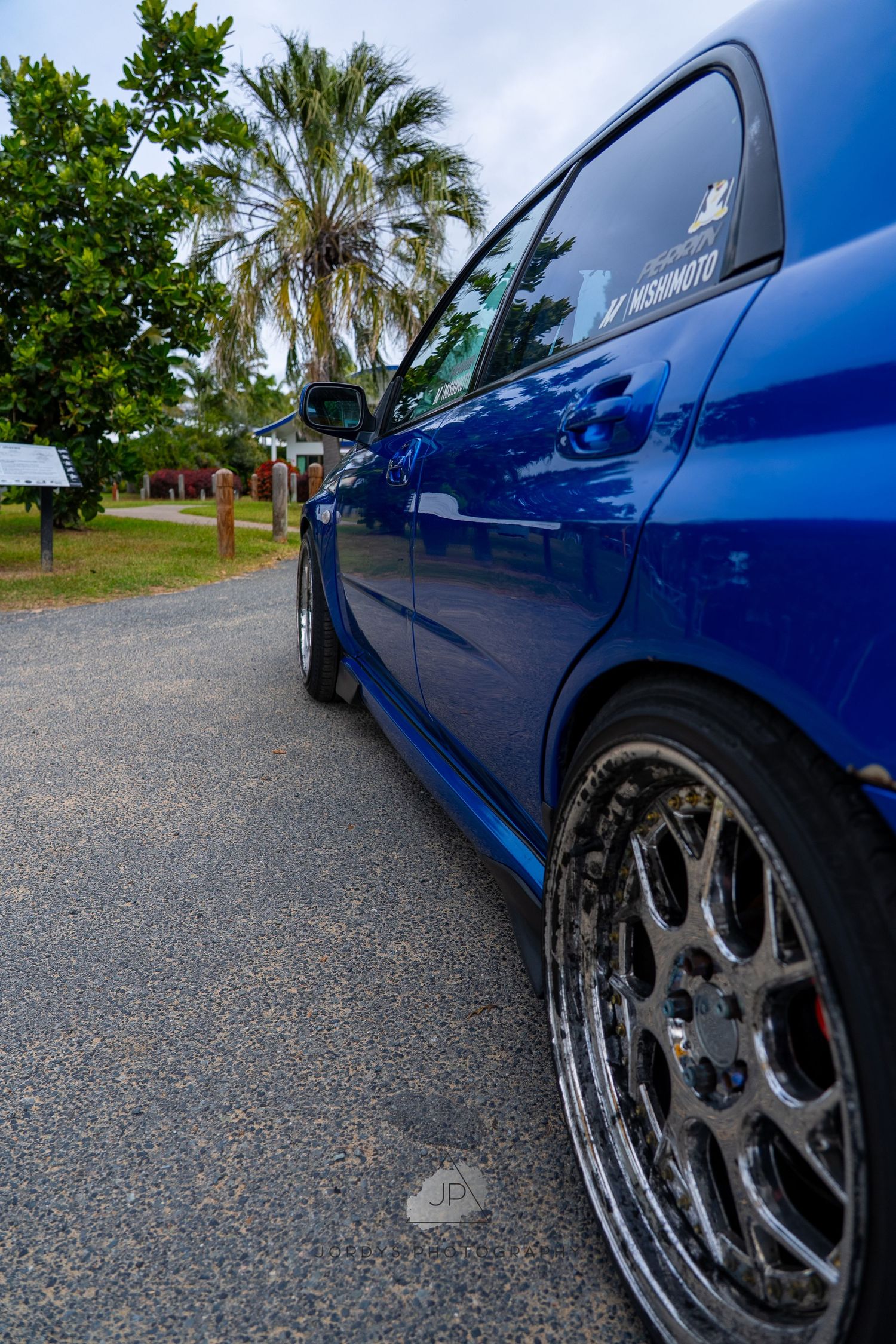 A series of five shots showing a blue car from different angles with custom wheels parked on gravel surface near palm trees.