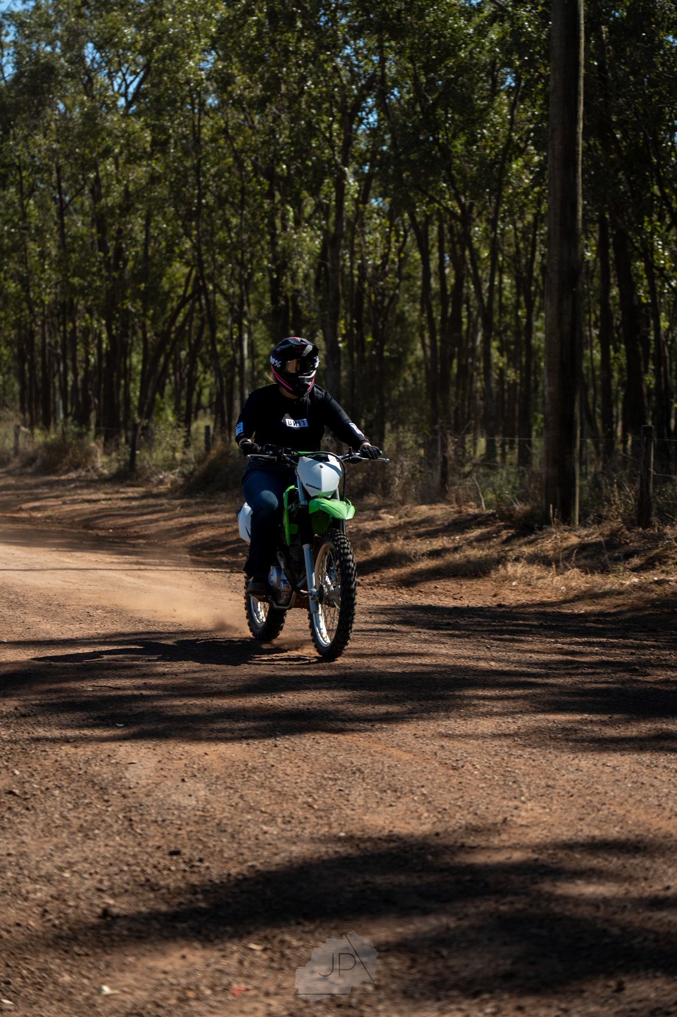 A dirt bike rider navigates through a wooded trail with sunlight filtering through the trees.