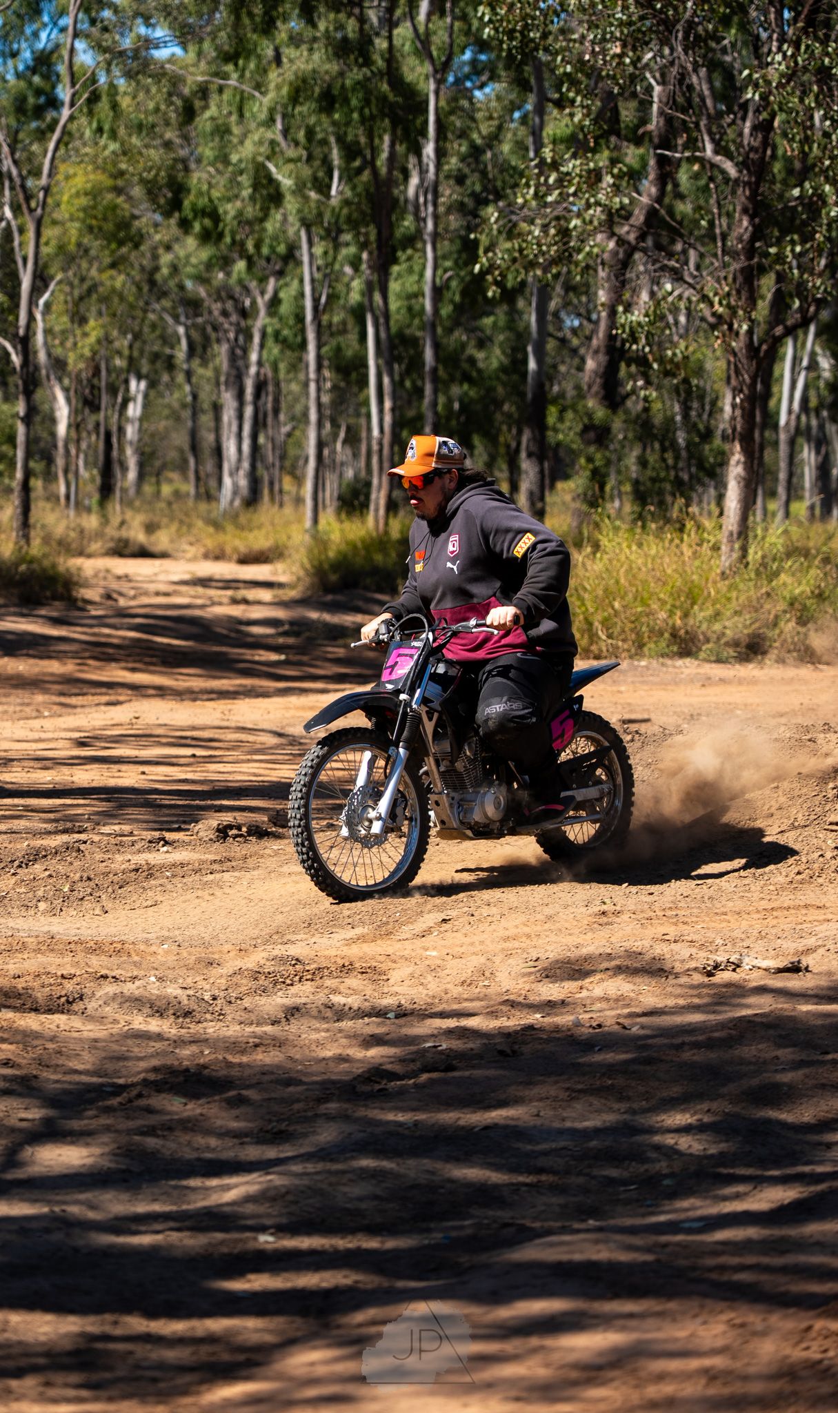 A dirt bike rider kicks up dust while navigating a trail through tall eucalyptus trees on a sunny day.