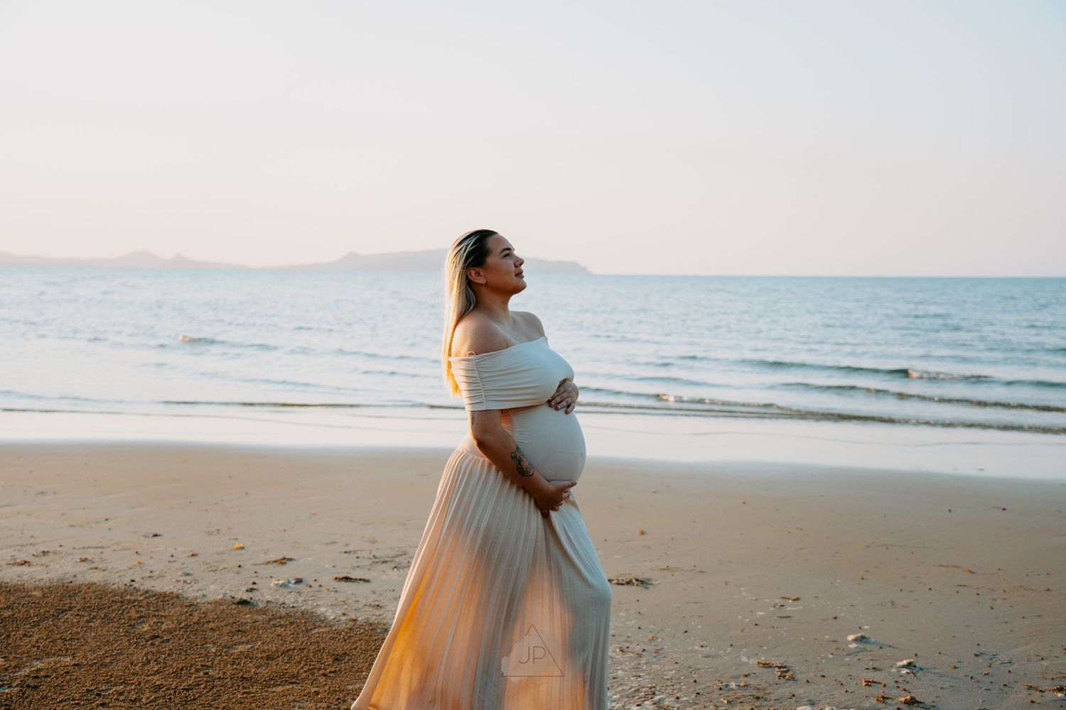 Maternity photoshoot on serene beach with woman in off-shoulder white gown at dusk.