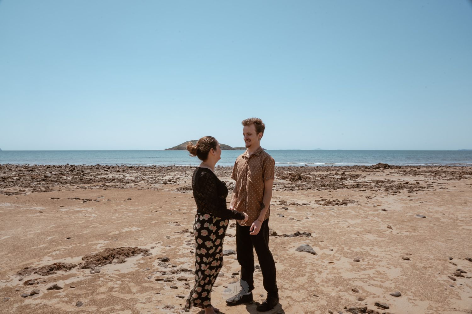 A couple explores the vast rocky desert landscape near a calm ocean under bright blue skies.