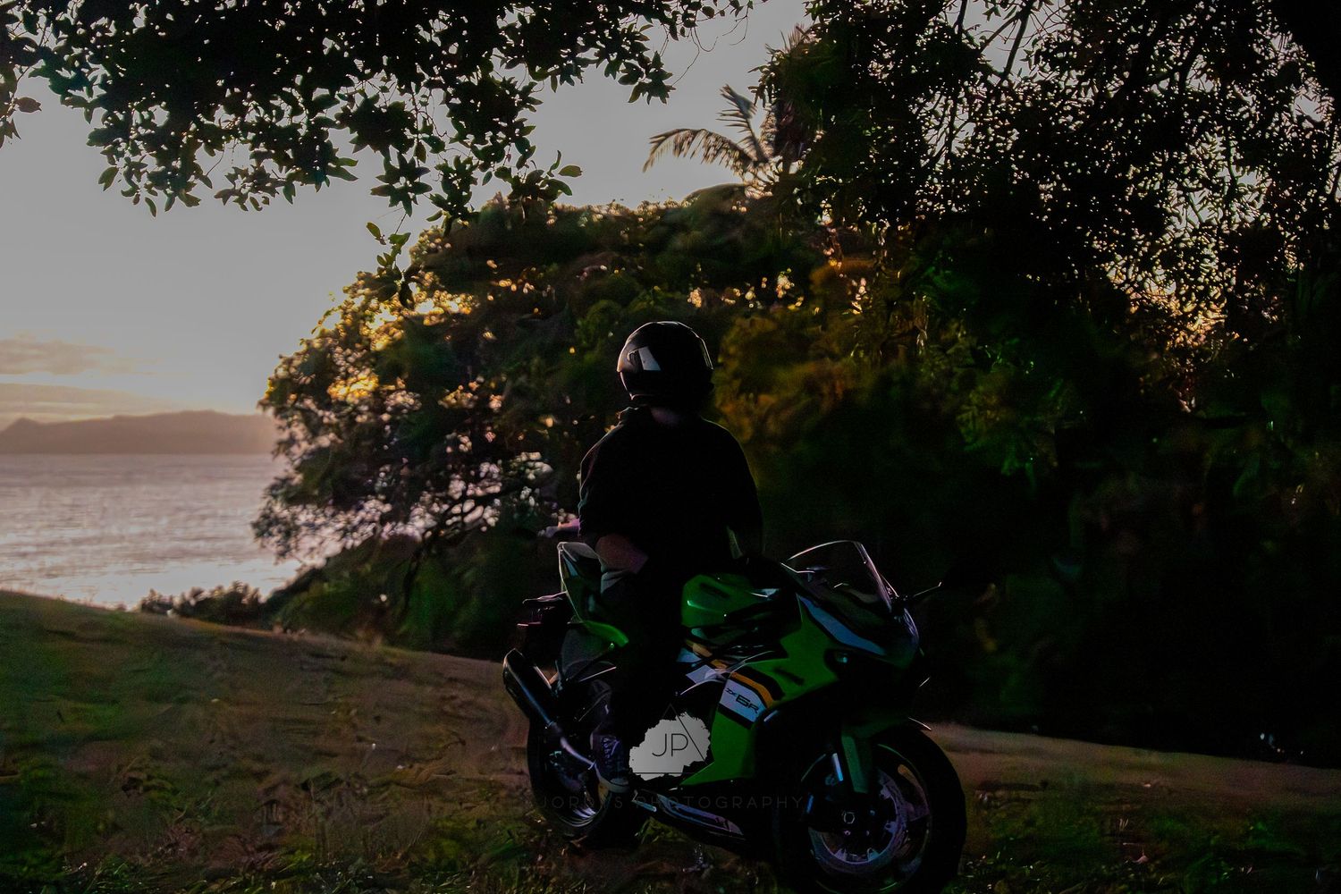 Six dark motorcycles parked in a row at sunset with trees and water in the background.