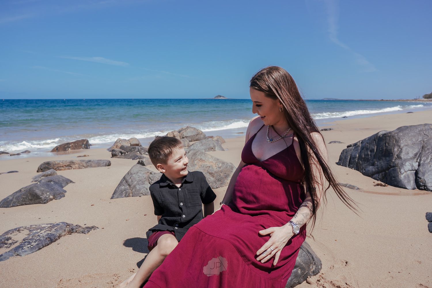 A heartwarming scene of a child and pregnant person sharing a moment on a rocky beach with blue ocean waves.