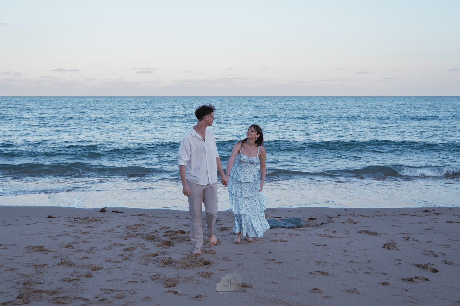 A couple in formal attire walks along a sandy beach at sunset with ocean waves in the background.