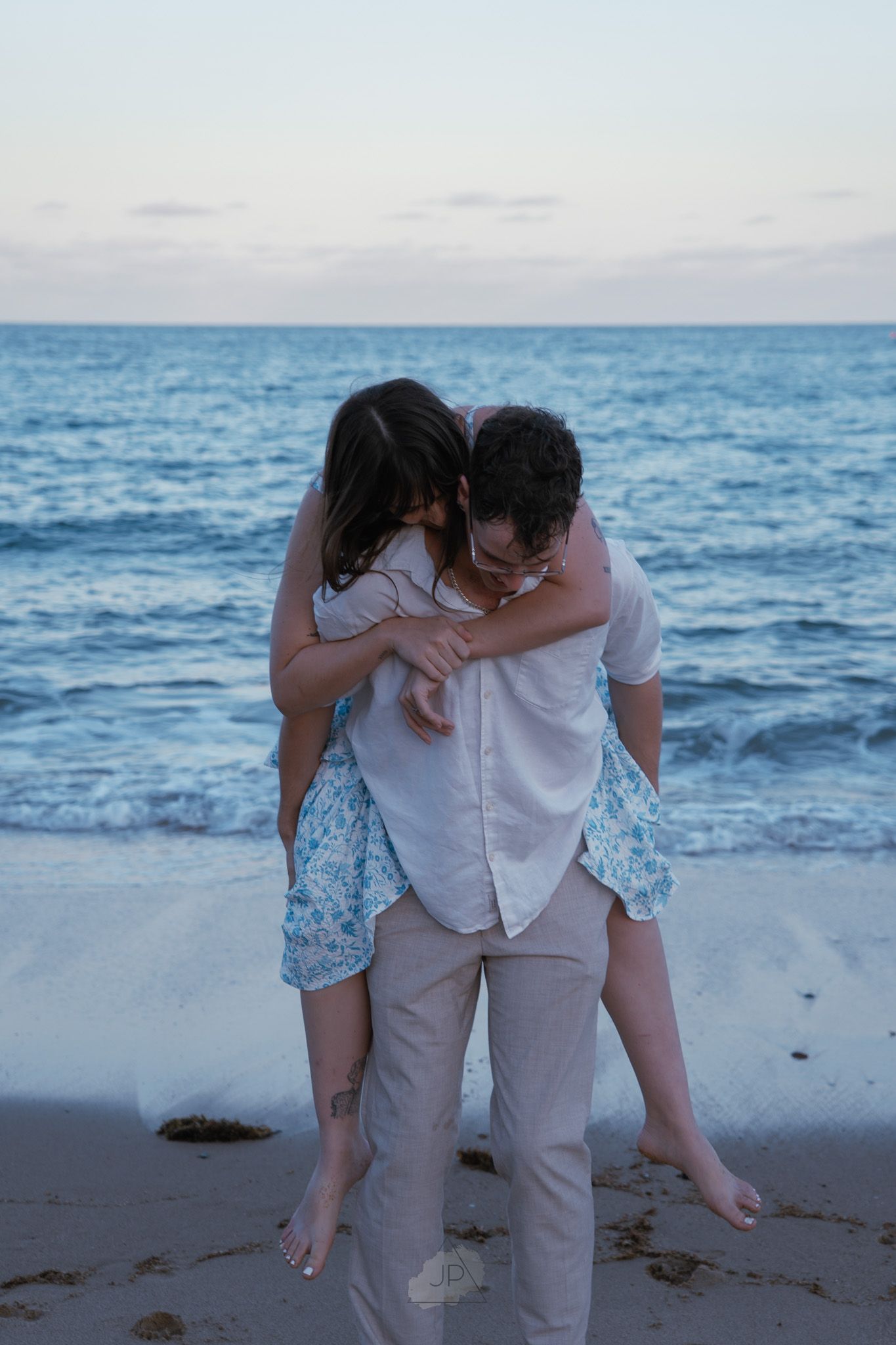 A couple embraces while walking along a beach at sunset with the ocean waves in the background.