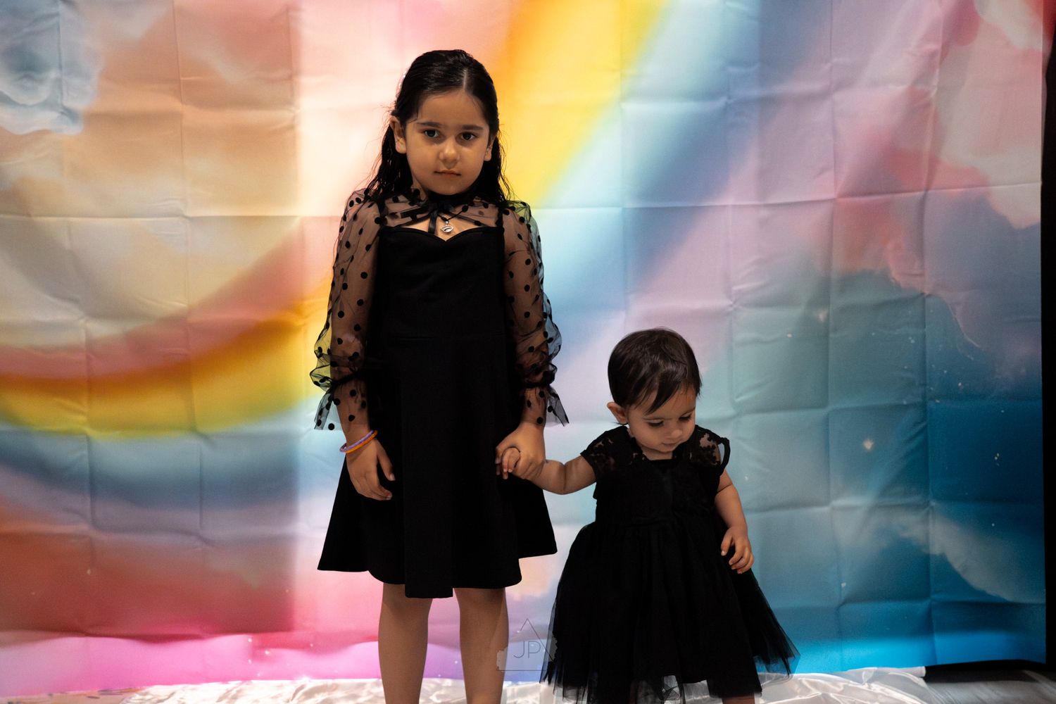 Two children in black dresses stand in front of a colorful rainbow backdrop.
