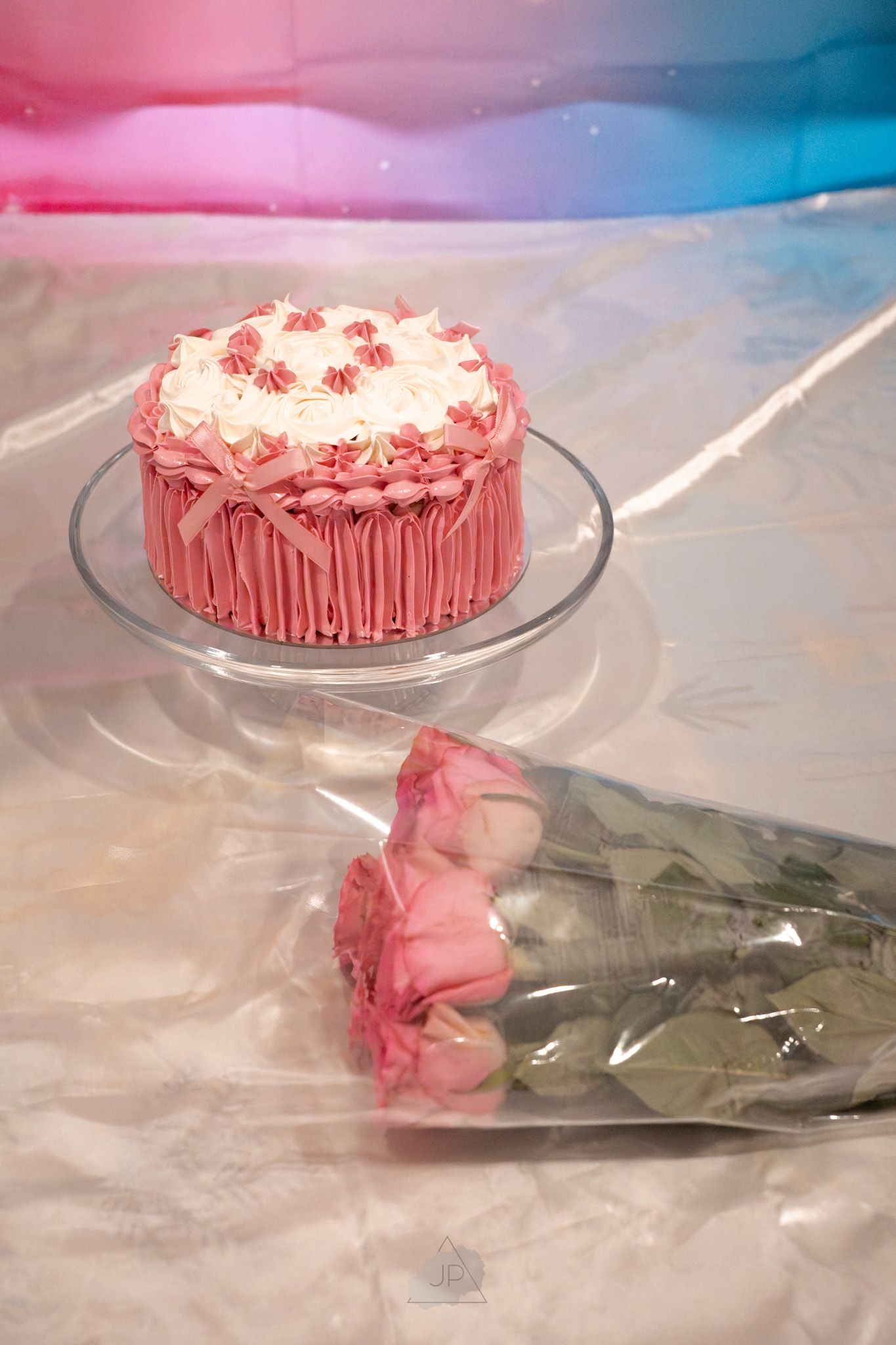 Pink frosted cake with delicate flower decorations displayed on a glass cake stand with roses in the background.