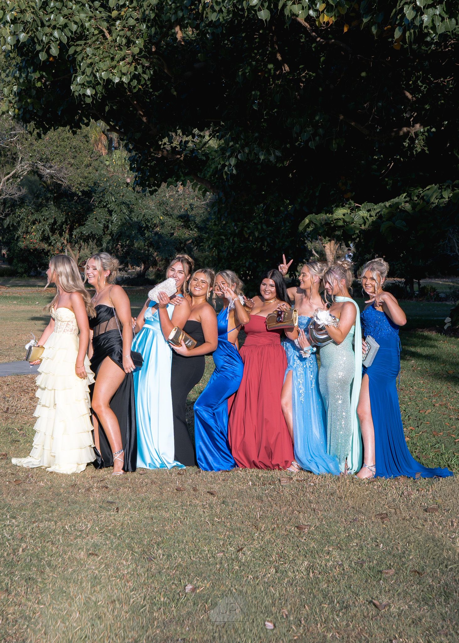 A group in coordinated blue and burgundy dresses poses playfully in a line on grassy ground during a sunny day.