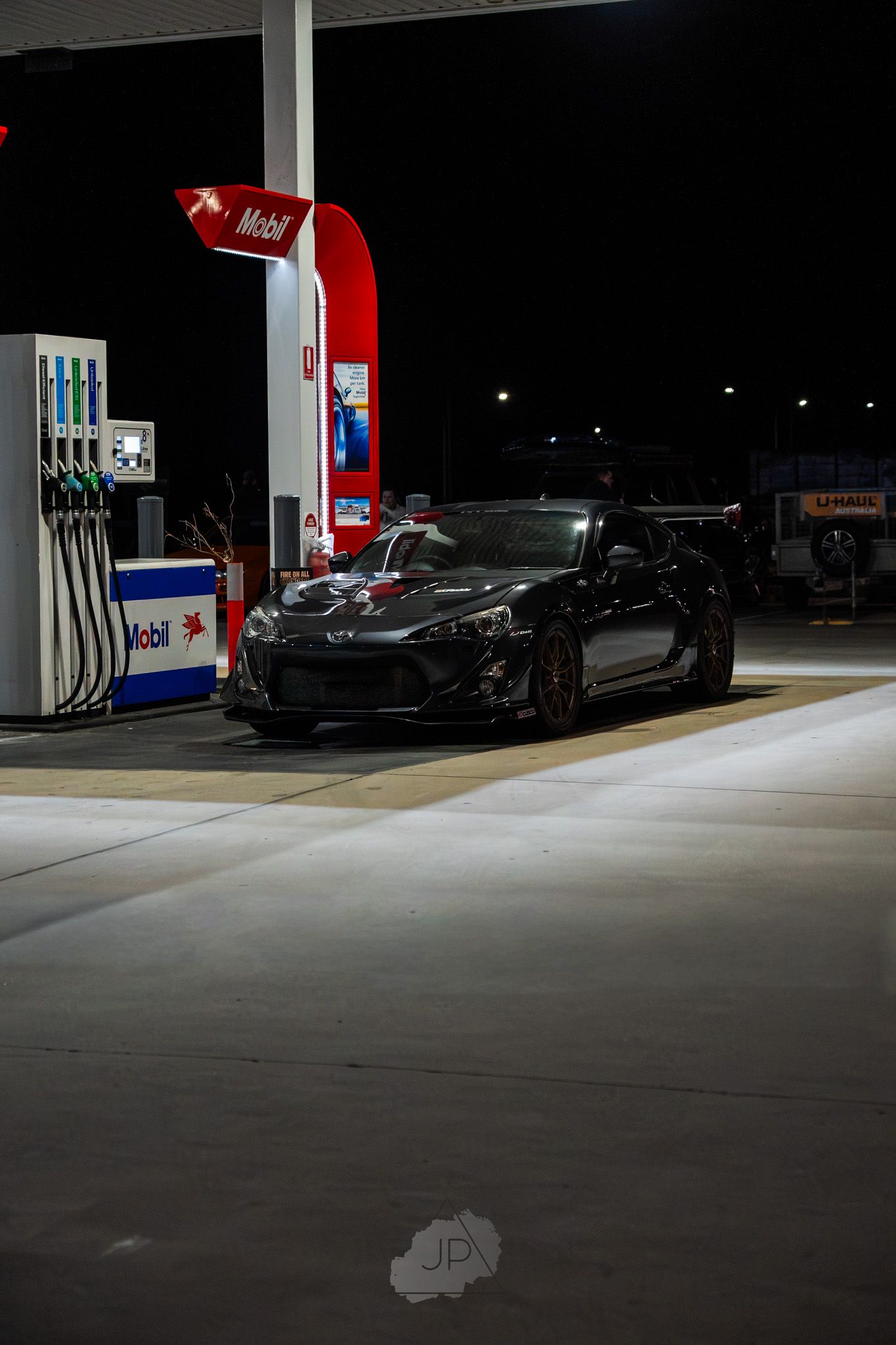 Dark gas station at night with fuel pumps and a black car parked under bright fluorescent lighting.