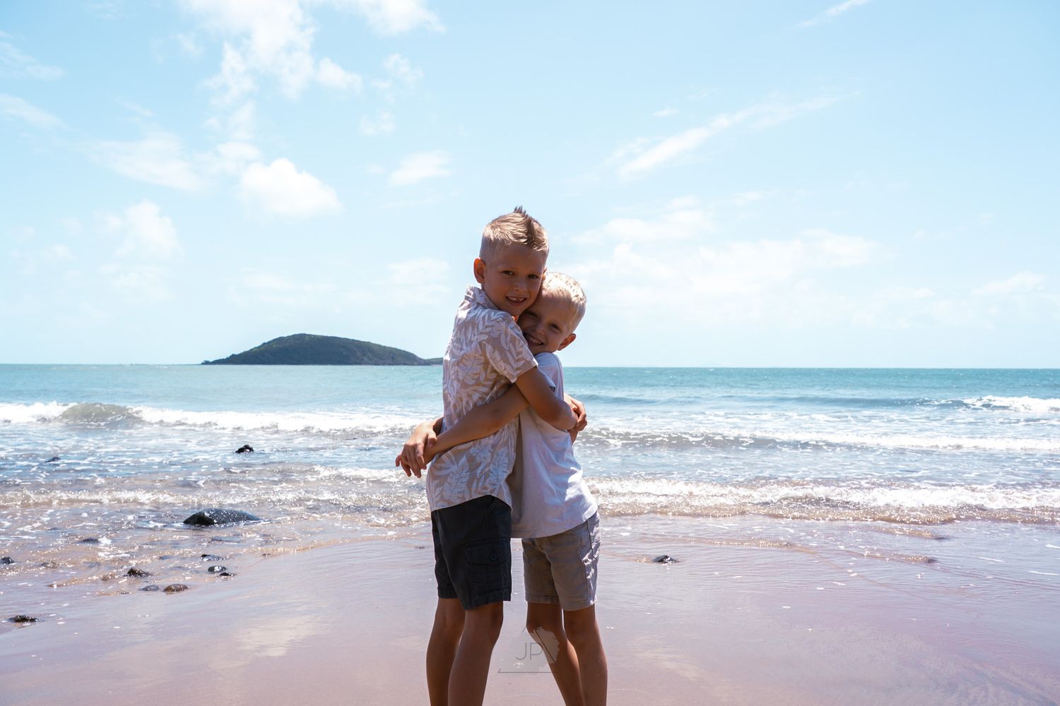 A couple embraces while standing on a sunny beach with waves and an island visible in the background.