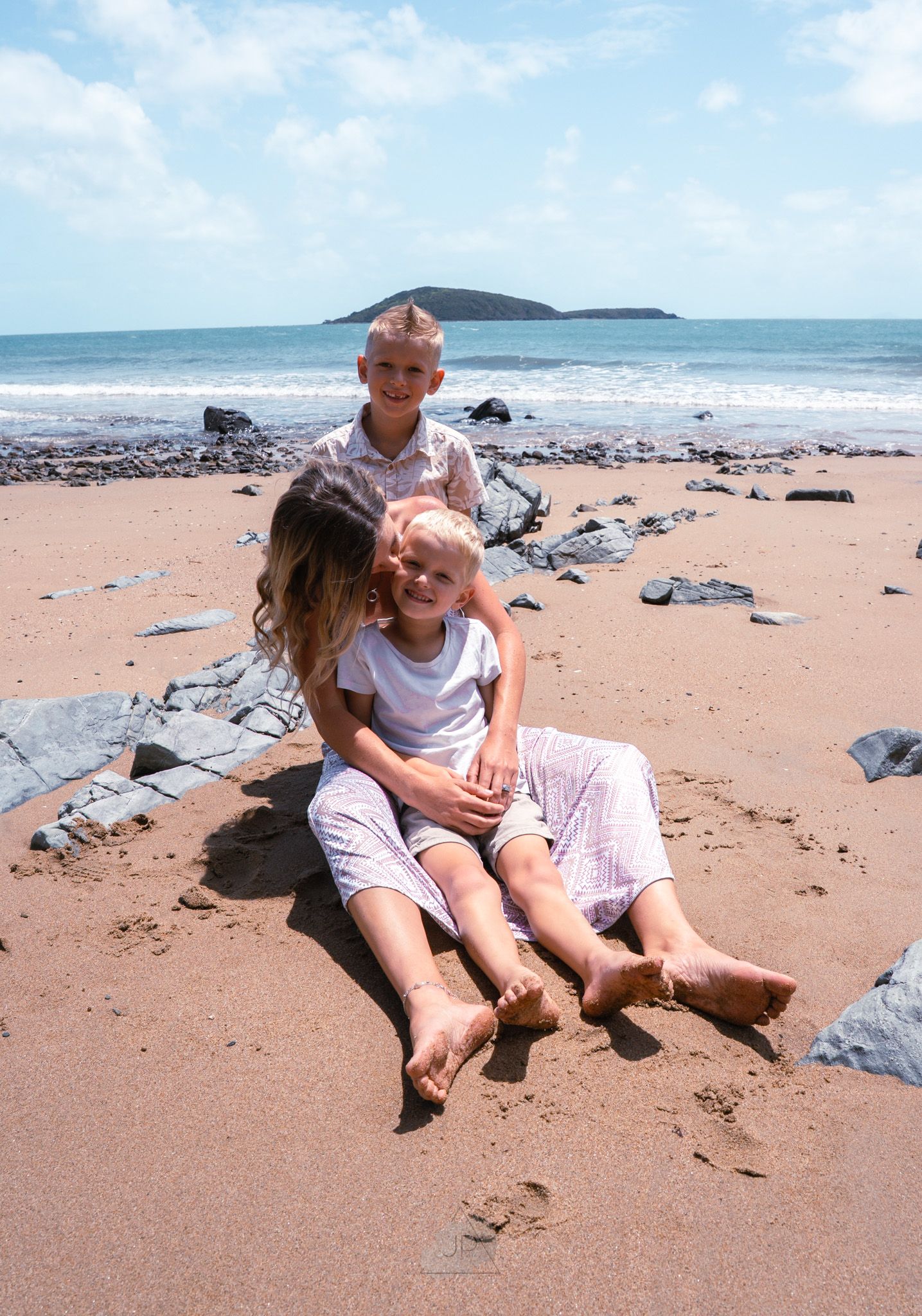 People relaxing on a sandy beach with rocks and an island visible in the ocean background on a sunny day.