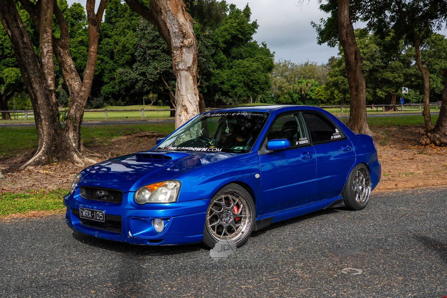 Blue Subaru Impreza WRX with custom wheels parked on a gravel path near trees.