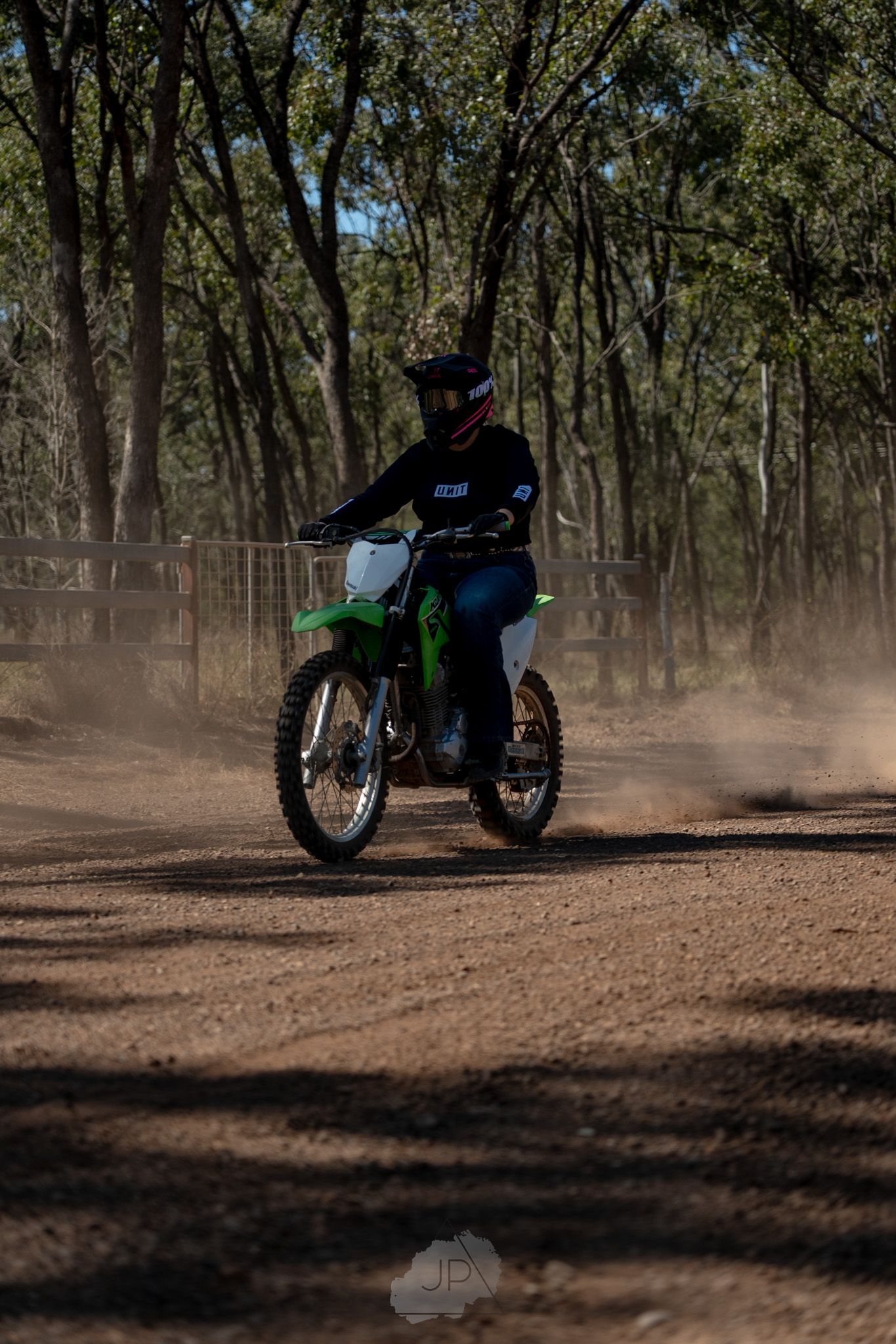 Dirt bike rider kicks up dust while riding through a wooded forest trail in dramatic lighting conditions.