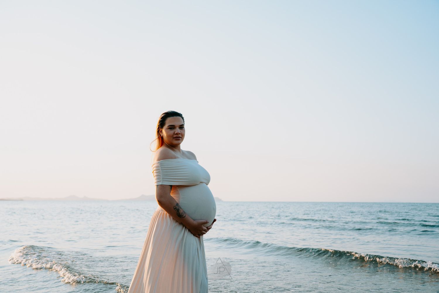 A person in a flowing white dress stands on a beach at sunset cradling their pregnant belly by the ocean waves.