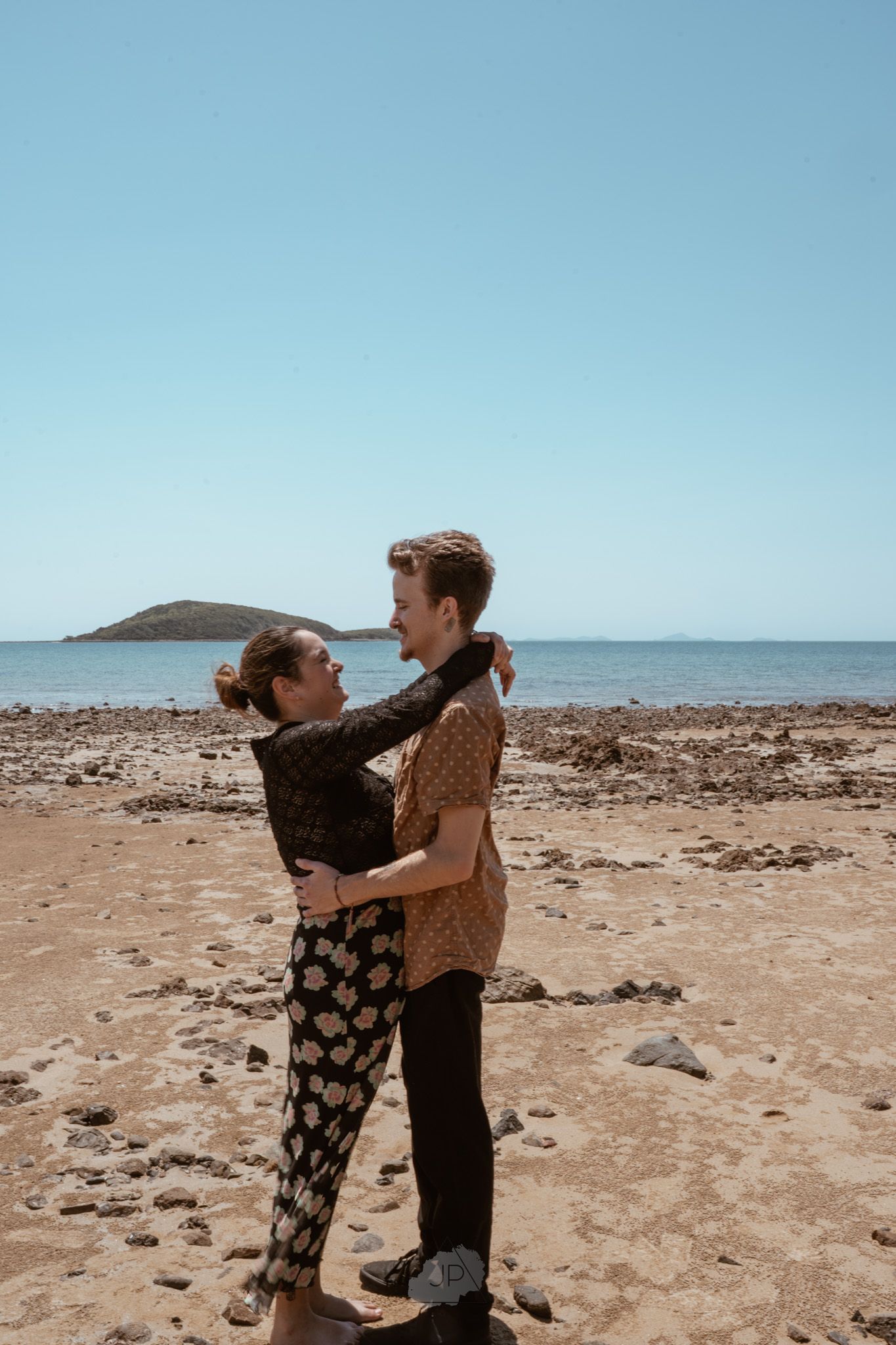 Two figures playfully interact on a barren rocky shore with ocean and mountains visible in the background.