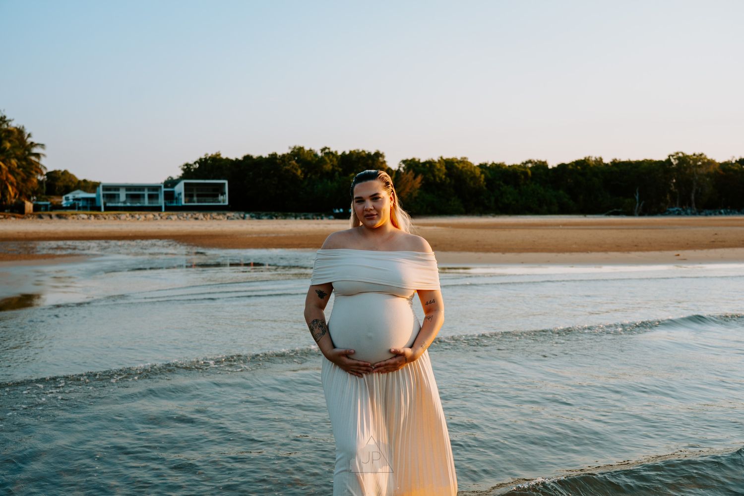 Woman in white dress walking along sandy beach at sunset holding bouquet of flowers.