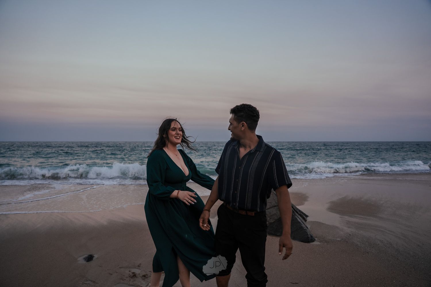 A couple shares a romantic moment at sunset on a sandy beach with ocean waves in the background.