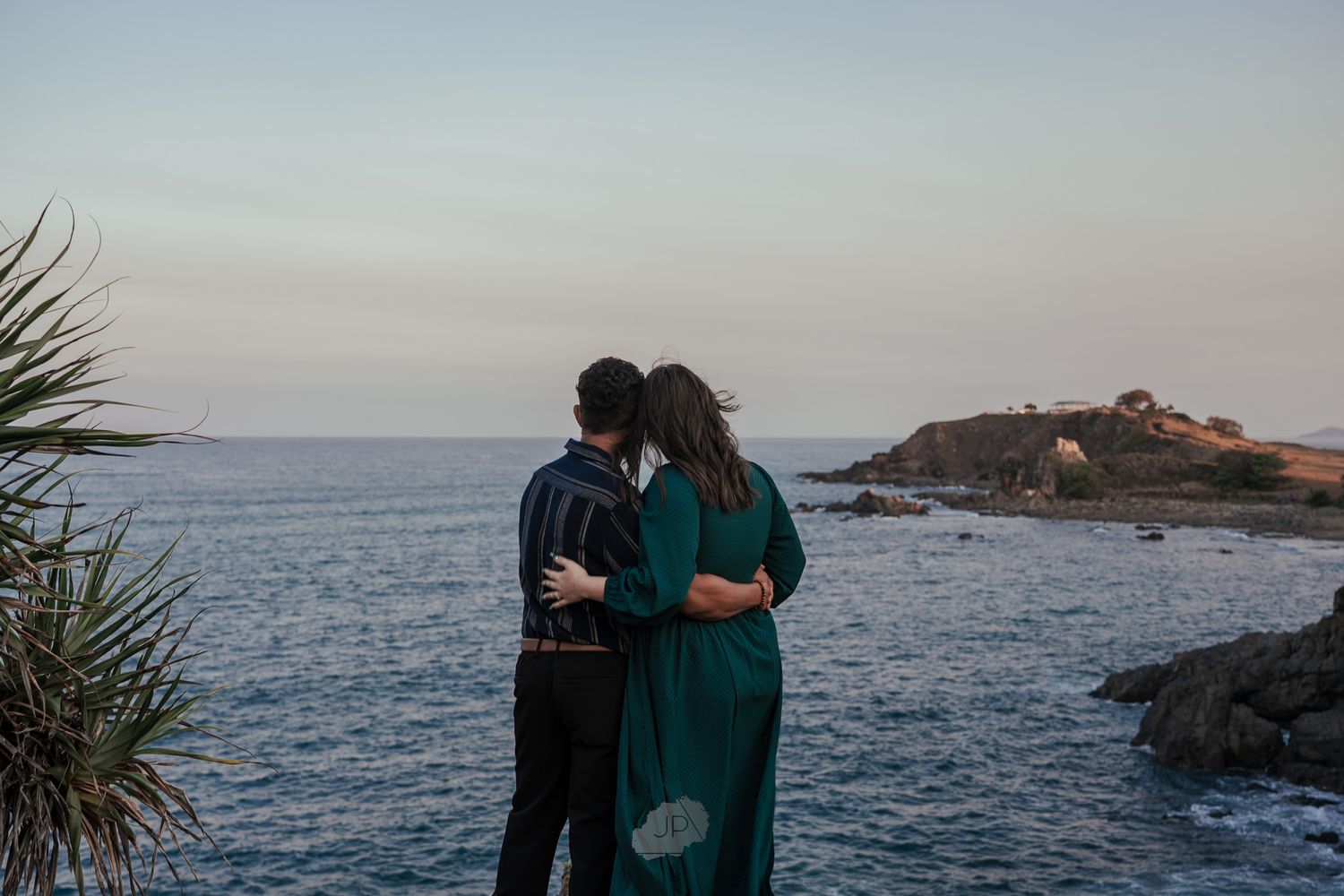 Two people embrace on a rugged oceanside cliff with waves crashing against the rocky shore below.