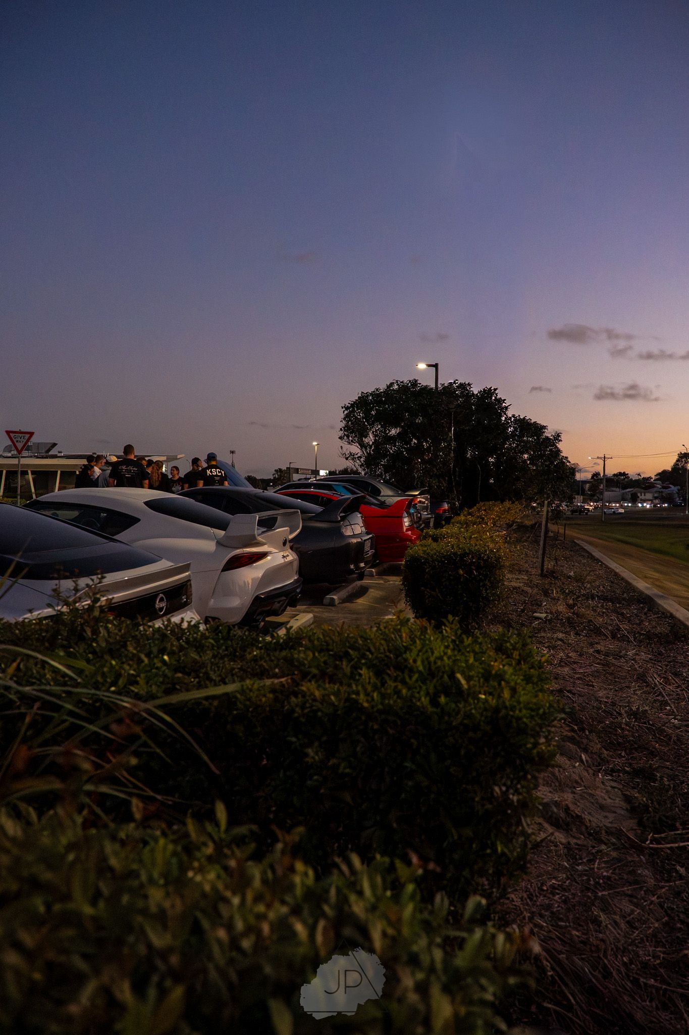 Cars parked in a lot at sunset with silhouetted trees and landscaping against a purple twilight sky.