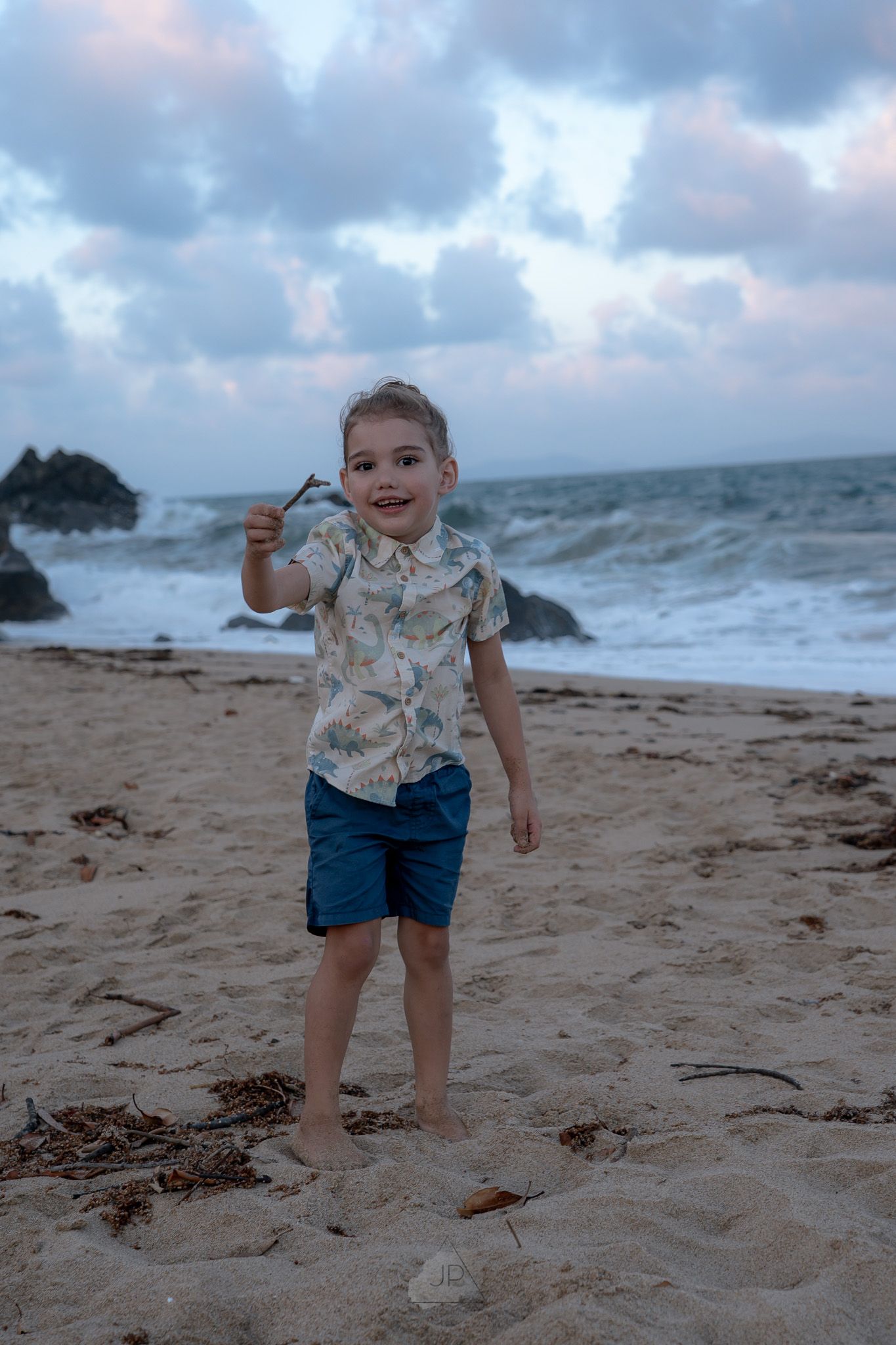A child explores a stormy beach at dusk, walking barefoot in the sand with waves crashing in the background.